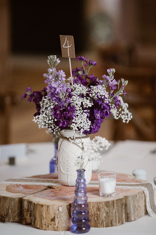 White mason jar centrepiece with purple and white flowers and a burlap table number.